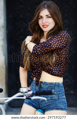 Lively young woman with long straight fair hair wearing on dark blouse and blue shorts is sitting on the bicycle and looking at camera on the street of old European city