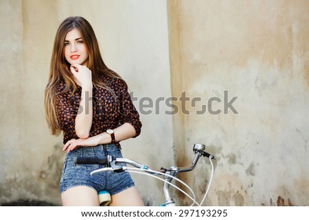 Lovely young woman with long straight fair hair wearing on dark blouse and blue shorts is sitting on the bicycle on the old wall background
