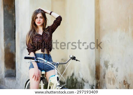 Young woman with long straight fair hair wearing on dark blouse and blue shorts is posing on the bicycle on the old wall background