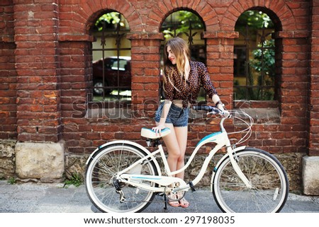 Happy girl with long fair hair wearing on dark blouse and  blue shorts standing with bicycle near the brick building, on the old city street