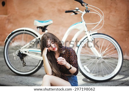 Young nice girl with long hair wearing on dark blouse and blue shorts sitting on tiled pavement and looking at her mobile phone and smiling with bicycle on a background on the street of old city