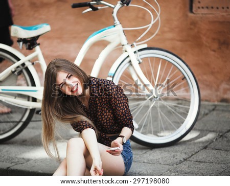 Beautiful young girl with long hair wearing on blouse and shorts sitting on tiled pavement, holding her mobile phone and have fun with bicycle on a background on the street of old city