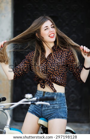 Young cheerful girl wearing on dark blouse and blue shorts with long straight fair hair is posing on the bicycle on the street of old city