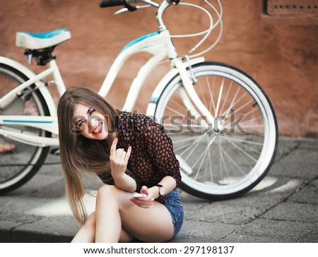 Nice young girl with long hair wearing on blouse and shorts sitting on tiled pavement, holding her mobile phone and laughing with bicycle on a background on the street of old city