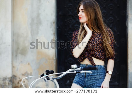 Attractive young girl wearing on dark blouse and blue shorts with long straight fair hair is sitting on the bicycle on the street of old European city