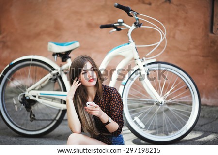 Young girl with long fair hair wearing on dark blouse and blue shorts sitting on tiled pavement and holding her mobile phone with bicycle on a background on the street of old European city