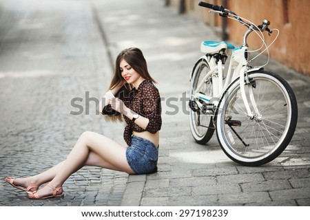 Smiling girl wearing on dark blouse and blue shorts with long straight hair sitting on tiled pavement near the bicycle and looking at her mobile phone on the street of old European city