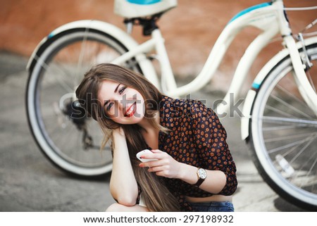 Smiling beautiful young girl with long straight fair hair wearing on dark blouse and shorts sitting on tiled pavement and holding her mobile phone with bicycle on a background on the street of city