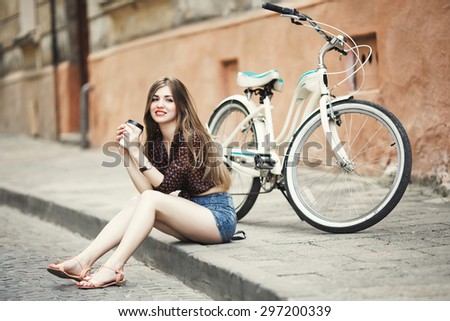 Young woman with long fair hair wearing on dark blouse and shorts is drinking coffee and smiling on tiled pavement with bicycle on a background on the street of old European city