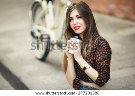 Pretty girl with long fair hair wearing on short dark blouse is drinking coffee on tiled pavement with bicycle on a background on the street of old city