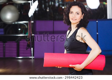 Waist up portrait of young active woman in sports wear holding pink yoga mat on a sports equipment background at the gym