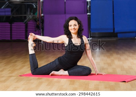 Smiling fitness woman with curly short hair wearing on black shirt and leggings make stretch on yoga pose on pink yoga mat on a sports equipment background at the gym