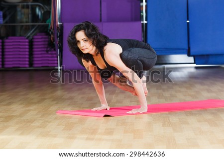 Young fitness woman with dark curly hair wearing on black shirt and leggings does yoga exercise on yoga mat on a sports equipment background at the gym