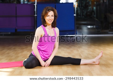 Smiling athletic girl with dark straight short hair wearing on violet shirt and black leggings is sitting on pink yoga mat on a sports equipment background at the gym