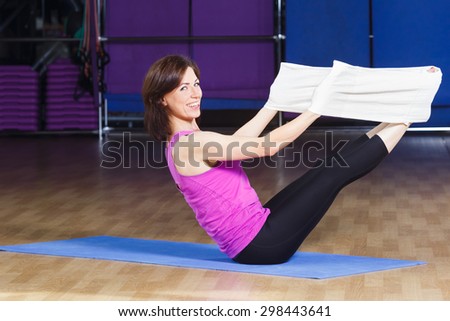 Beautiful fitness woman with dark hair wearing on violet shirt and black leggings is training with a white core band on a sports equipment background at the gym
