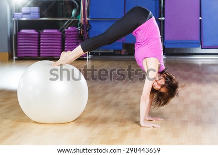 Young fitness woman with short dark hair wearing on violet shirt and black leggings does exercises and smiling on a white fitball on a sports equipment background at the gym