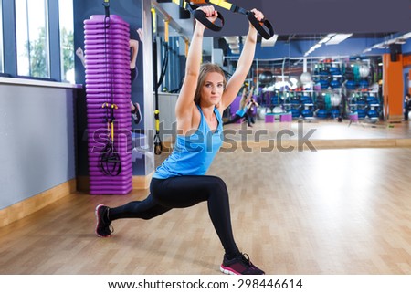Beautiful brunet fitness girl wearing in blue shirt is crouching with TRX in the gym