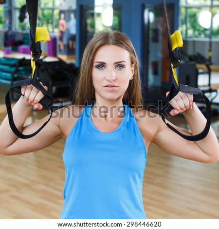 Cute brunet fitness girl with straight brunet hair wearing in blue shirt is doing exercises with TRX in the gym