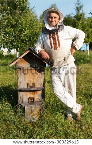Happy beekeeper in protective hat and white shirt, is standing near the wooden beehive in the bee-garden with multicolor wooden beehives on the background, close up