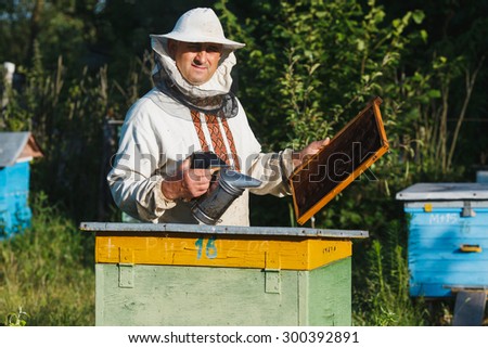 Apiarist in a protective hat and white shirt making inspection in apiary and looking at the camera in the garden