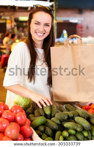 Pretty young woman with paper bag is standing near the boxes with tomatoes and cucumbers and buying her vegetables, on the market, waist up