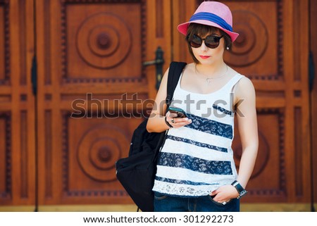 Pretty young woman using smartphone, with black backpack on shoulder, wearing sunglasses, stylish pink hat and striped shirt and denim shorts. On old wooden doors with a pattern background. Copy space