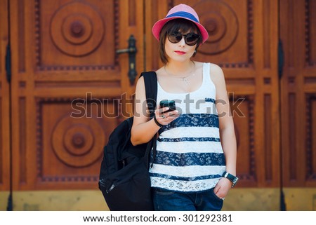 Pretty young woman with smartphone and black backpack on shoulder, wearing sunglasses, stylish pink hat and striped shirt and denim shorts. On old wooden doors with a pattern background. Copy space.
