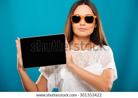Cute young girl, with long hair, wearing in white blouse, and orange sunglasses, is holding tablet on blue background, waist up