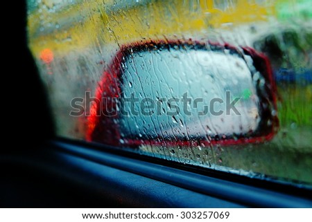 Rain drops rolling on window, and a red car side view mirror on a background, close up