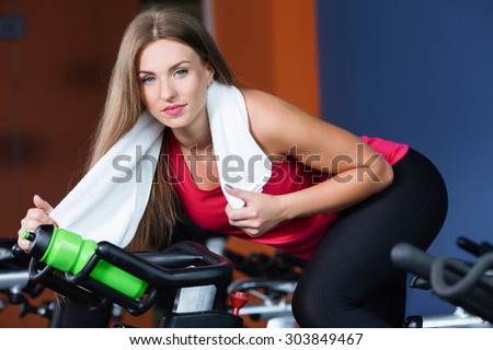 Pretty brunette woman is posing on exercise bike with bottle of water and white towel on her neck, in the gym, waist up