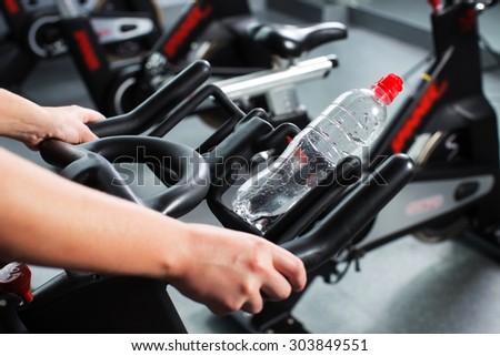Cropped image of woman\'s hands holding exercise bike, with bottle of water on it, on the gym, close up