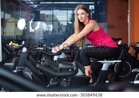 Attractive girl, wearing in pink shirt and black leggings, doing indoor biking exercise, with glass on the background, full body