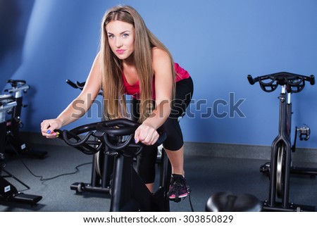 Young woman, wearing in pink shirt and black leggings, doing indoor biking exercise, on blue wall background, full body