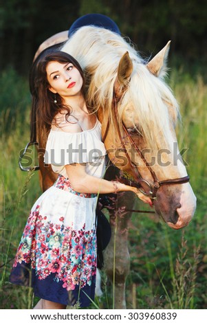 Young cute woman in white short dress, standing with brown strong and muscular horse in green summer field, waist up