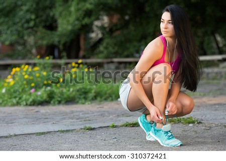 Full length portrait of active jogging female runner, with long dark hair, preparing shoes for training and working out at fitness park