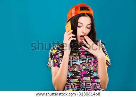 Pretty girl, with dark straight hair, wearing in colorful blouse and orange cap, talking on her smart phone on blue background, waist up