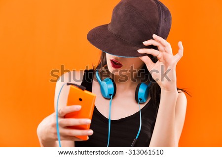 Charming young woman, in black blouse and cap, holding smart phone in her hand and blue headphones hanging on her neck - isolated on orange background, in studio, waist up