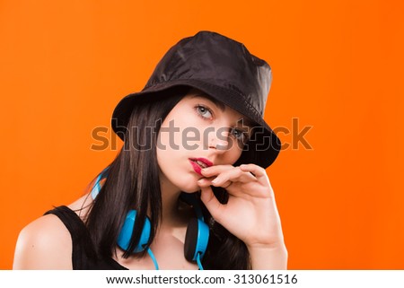 Nice young girl, with blue headphones on her neck, wearing in black blouse and cap, posing on orange background, in studio, waist up