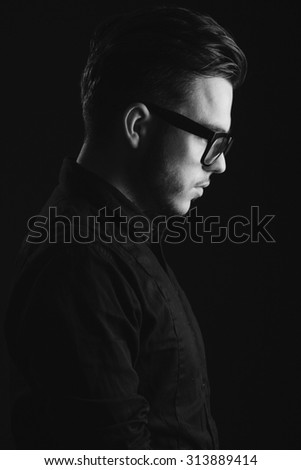 Side view image of a young handsome man with beard, wearing on black shirt and glasses, on dark studio background, close up
