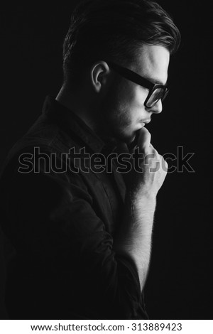 Side view of attractive brunette man with beard, wearing on black shirt and glasses, looking down, on dark studio background, close up