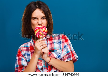 Angry young girl, with short hair, wearing in red checkered shirt, posing with colored candy, on blue background in studio, waist up