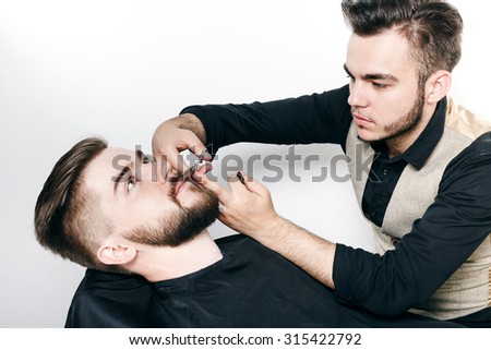 Young barber, wearing in black shirt and waistcoat, cutting bearded brunette customer with scissors, on gray background, in studio, waist up