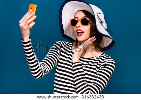 Charming brunette woman, wearing in striped blouse and white beach hat, is taking selfie with her smart phone, on blue background, in studio, waist up