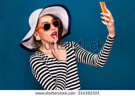 Coquettish young woman, wearing in striped blouse, orange sunglasses and white beach hat, is taking selfie with her smart phone, on blue background, in studio, waist up