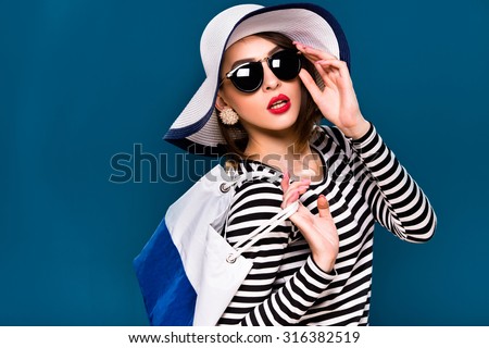 Nice brunette woman, with short haircut, wearing in striped blouse, black sunglasses and white hat, is posing with white and blue bag, on blue background, in studio, waist up