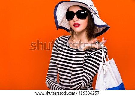Beautiful brunette girl, with short haircut, wearing in striped blouse, black sunglasses and white hat, is posing with white and blue bag, on orange background, in studio, waist up