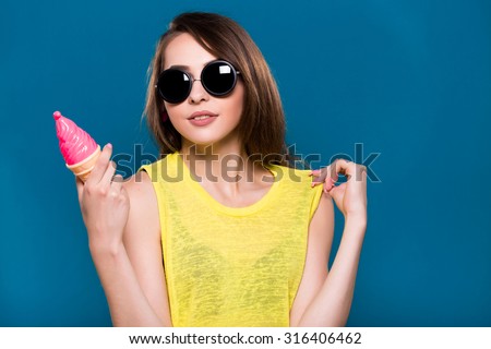 Charming young girl, wearing in yellow blouse and black sunglasses, is posing with ice cream in her hand, on blue background, waist up