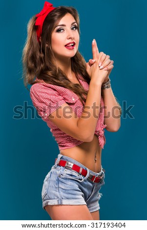 Cheerful pretty girl, with piercing in the navel, wearing in retro checkered shirt, shorts and red kerchief on her head, having fun on blue background in studio, waist up