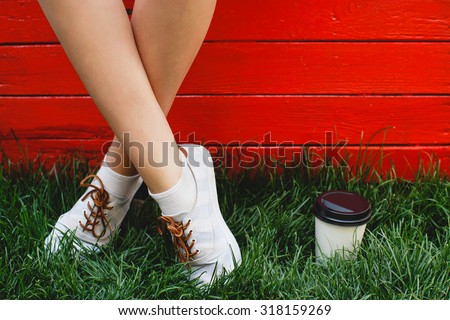 Feet of young girl, in white socks and sneakers, standing on green grass near the cup of coffee, on red wooden background, close up