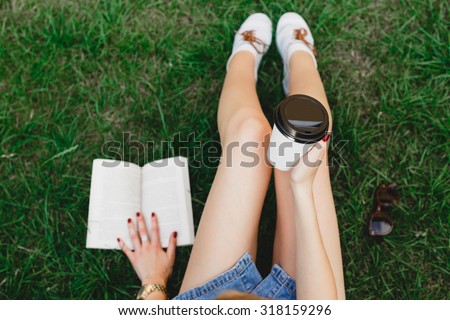 Slim girl, wearing in blue shorts and sneakers, sitting on the green grass with cup of coffee and book, sunglasses lying on the grass, close up. Point of view